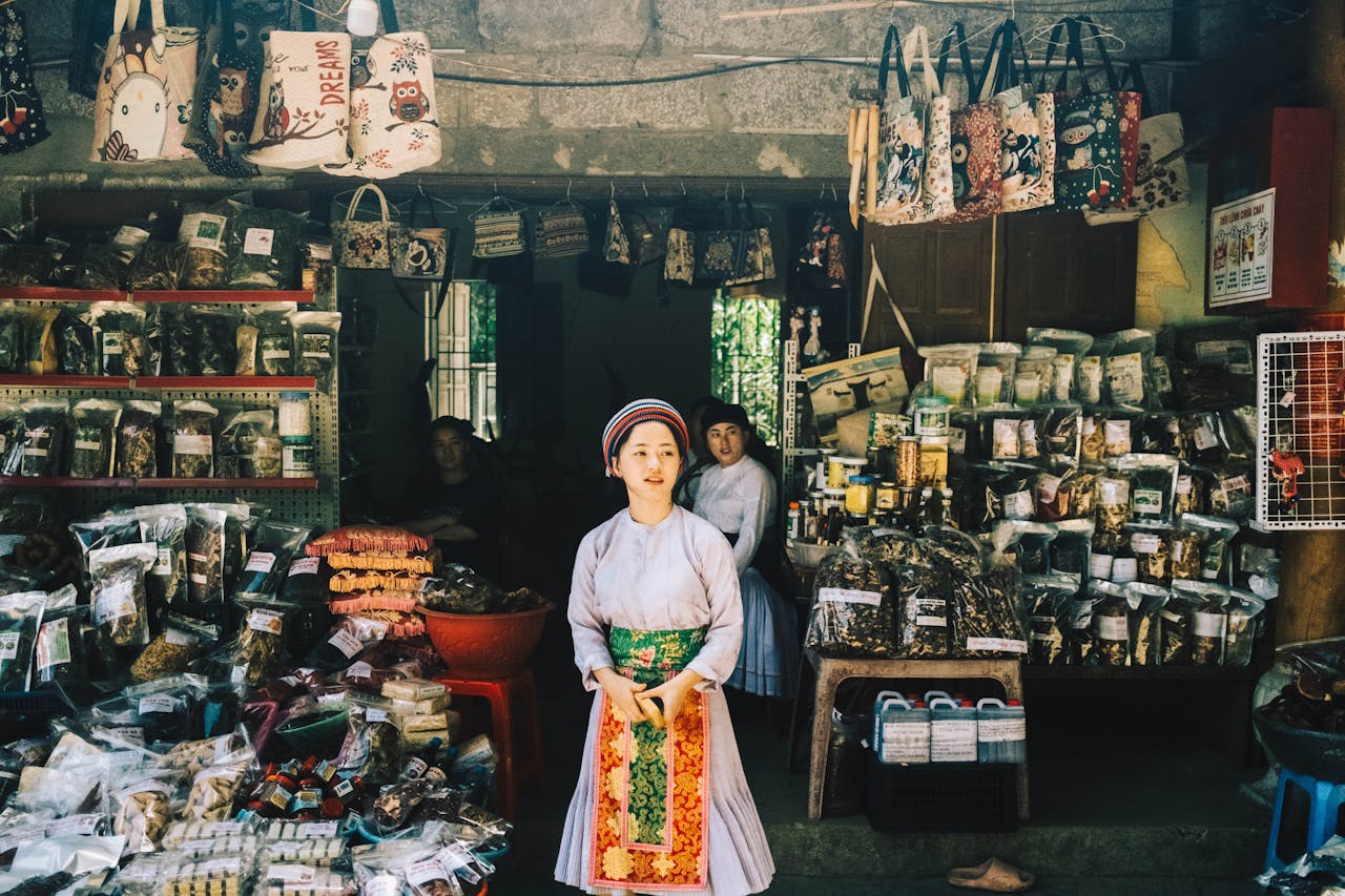 A woman in traditional attire stands in front of a vibrant market stall showcasing various goods and crafts.